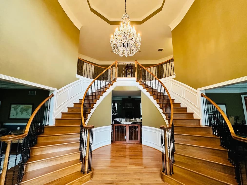 This is an image from Painting Contractors of MD, located in Maryland. The interior view features a grand two-sided curved staircase with wood steps, white wainscoting, and a large chandelier overhead.