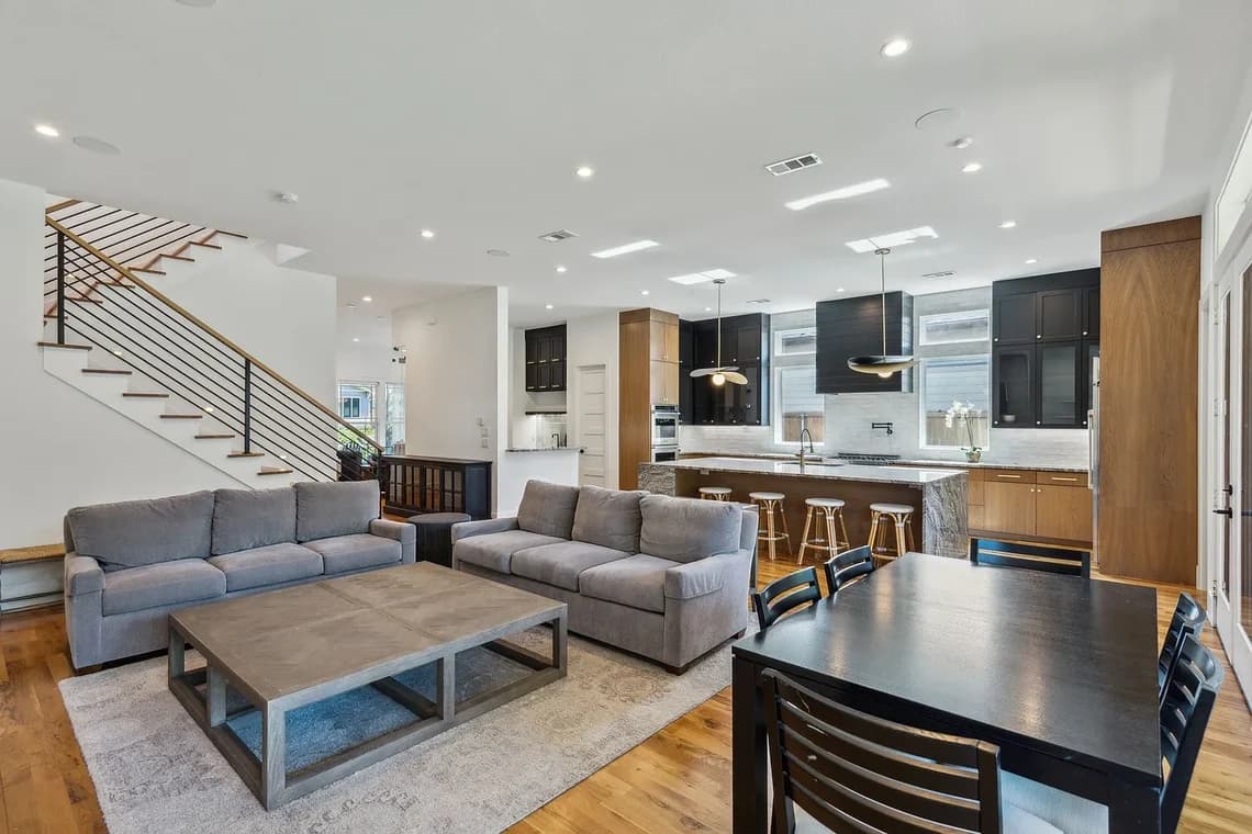 "This is an image from Highland Painting Co., located in Texas. The photo shows an open-concept living area with gray sofas, a wood dining table, and a modern kitchen featuring dark cabinetry and natural wood accents."