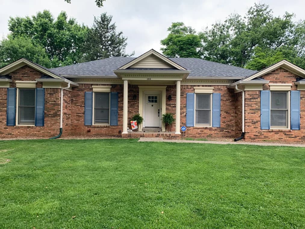 This is an image from Serious Business Painting LLC, located in Kentucky. The image shows a single-story brick home exterior with blue shutters, white trim, and a freshly maintained front facade.