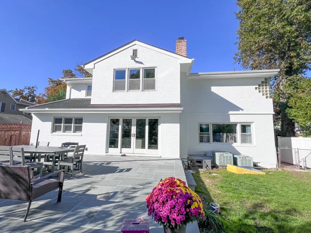 This is an image from Elite Painting Services Plus, located in New York. The backyard view highlights a freshly painted white brick home with a large patio area, outdoor seating, and vibrant garden flowers.