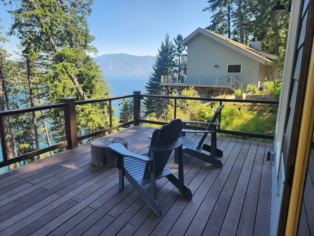 This is an image from Honey Do Painting LLC company, located in Bonners Ferry, ID The image is a lakeside view from a freshly stained wooden deck with Adirondack chairs and log table, surrounded by pine trees and overlooking a hillside home and blue water, project by Honey Do Painting