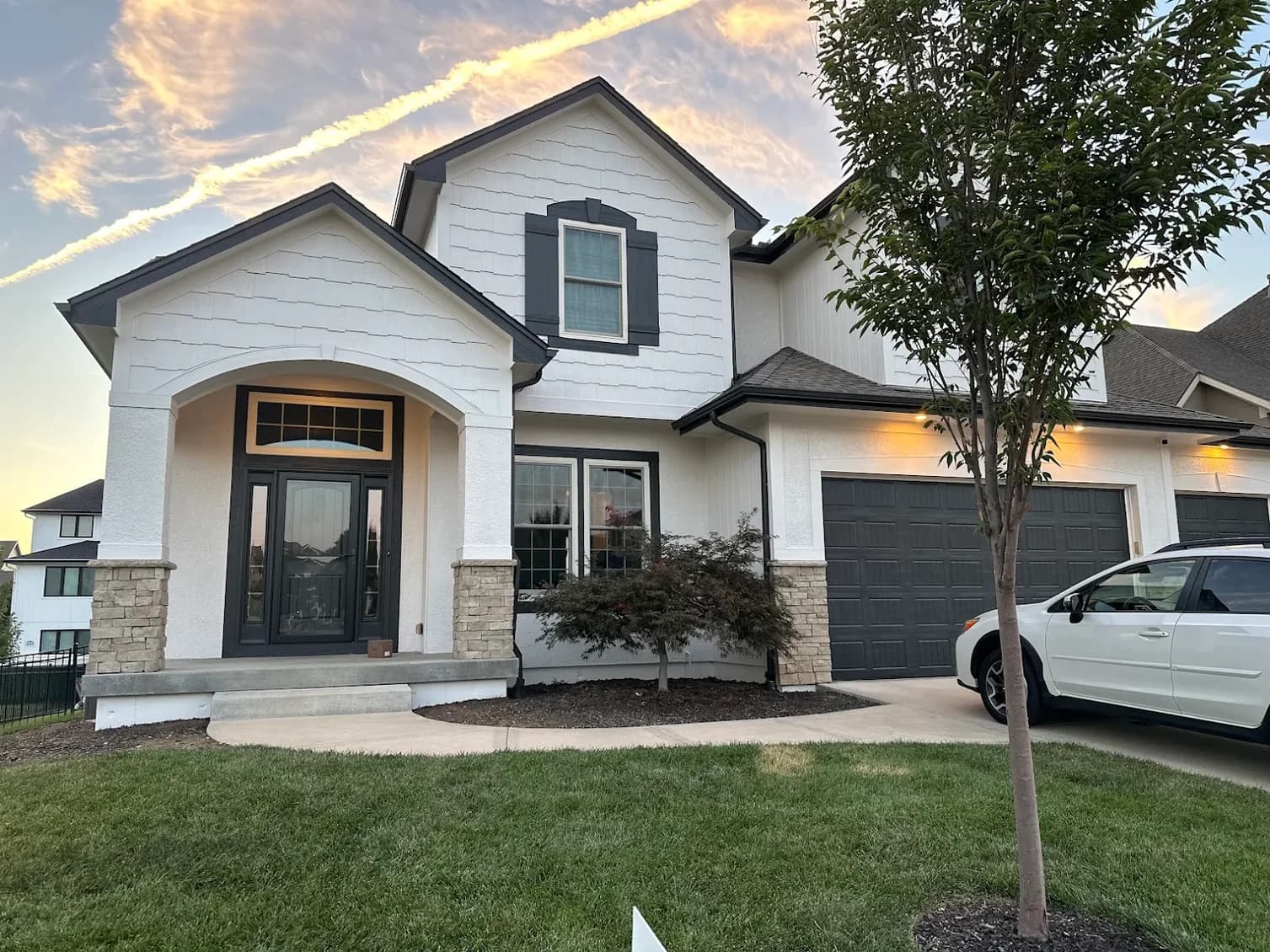This is an image from Mission Painting and Home Improvements. The image shows a modern two-story home with newly painted white stucco, dark gray trim, and matching garage doors, showcasing exterior painting results.