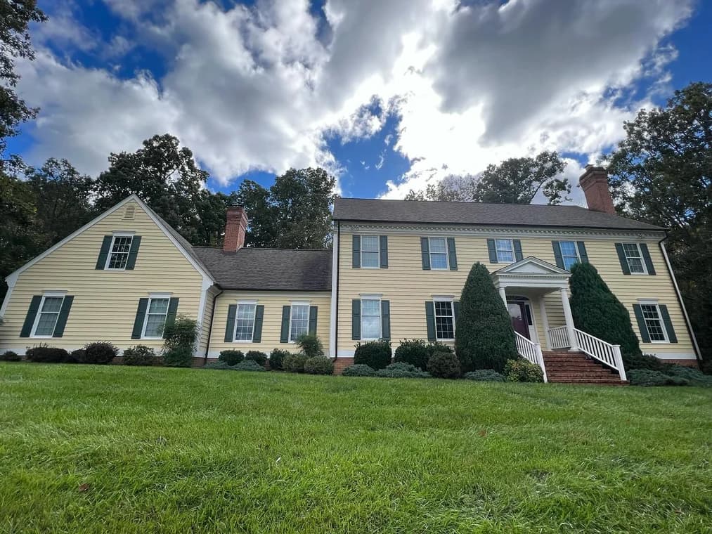 This is an image from Paint Perfection, located in Maryland. The photo shows a large yellow-sided two-story home with green shutters and white trim, freshly painted and surrounded by trees and a wide grassy lawn.