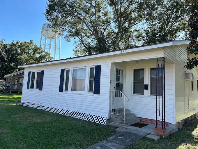 This is an image from 360 Painting of Lafayette, located in Louisiana. The photo shows a single-story white home with dark blue shutters, a small front porch, and a nearby water tower visible against a clear blue sky.