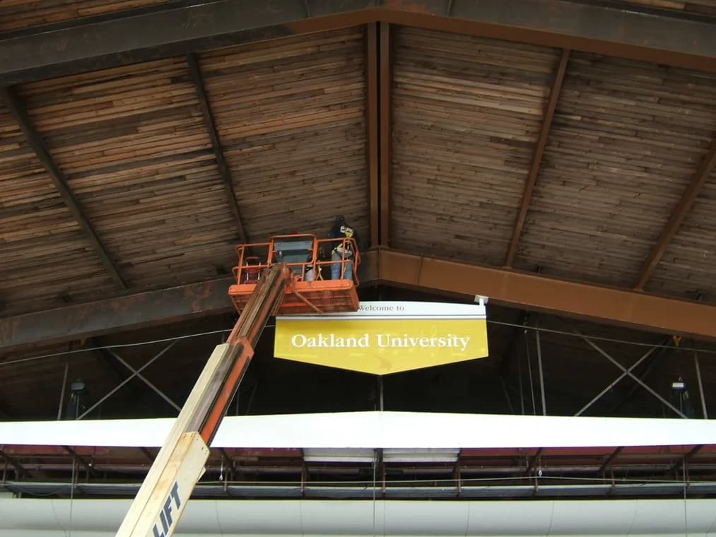 This is an image from The Exterior Painting Experts - F&H Painting company, located in Oxford, MI The image is a F&H Painting crew performing high-reach interior ceiling painting at Oakland University using a boom lift under a large wooden beam structure.