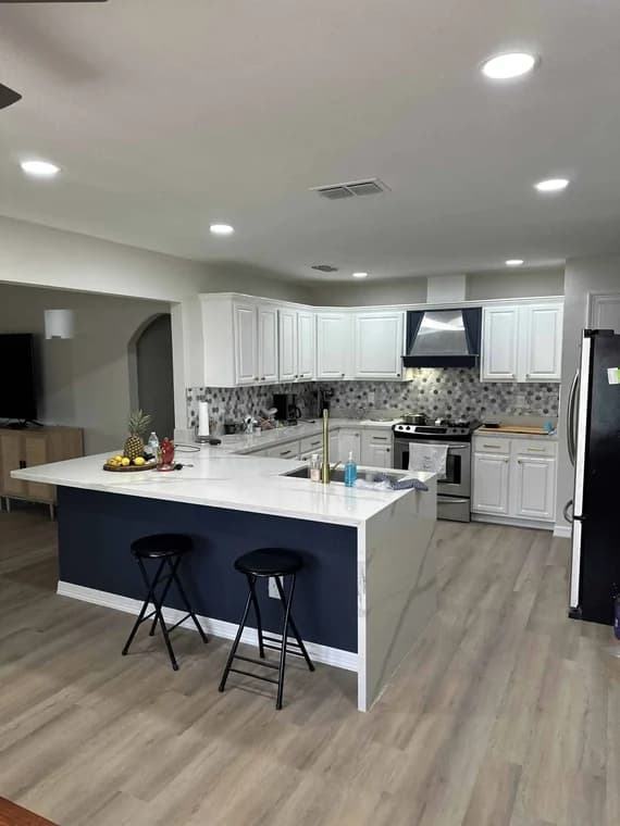 "This is an image from Armored Painting, located in Texas. The kitchen features freshly painted white cabinets, a navy blue island base, modern backsplash tile, and updated lighting over a light wood floor."