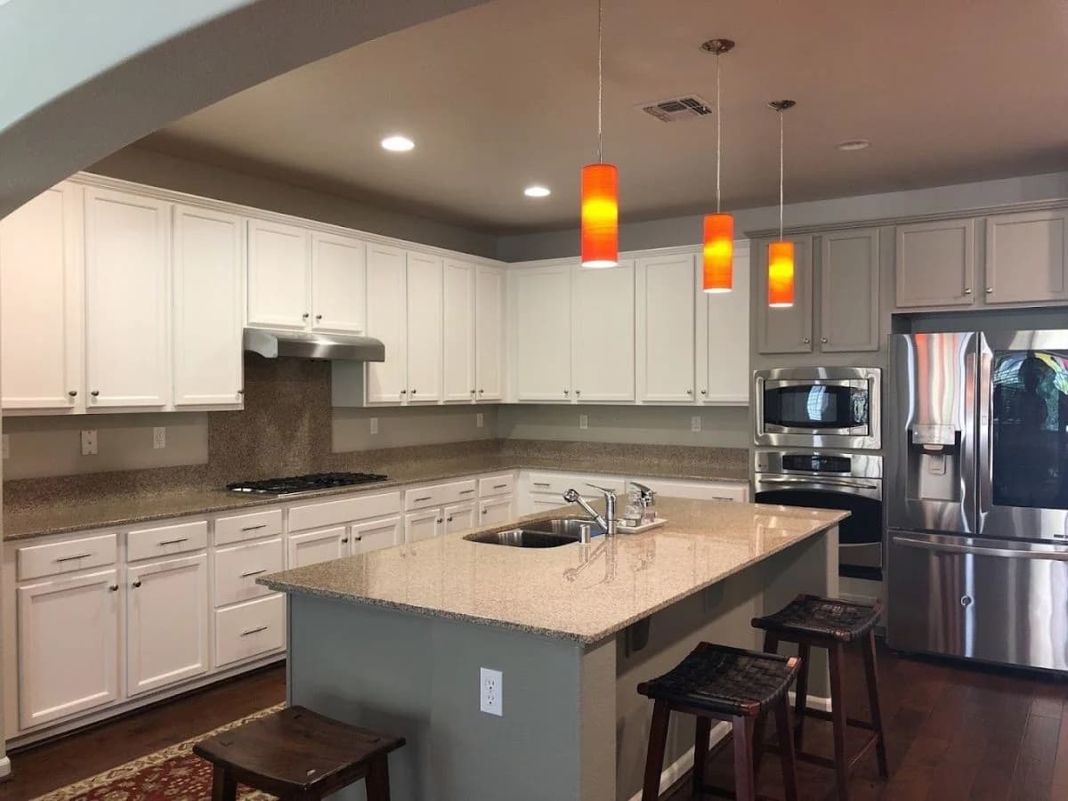 This is an image from Brooks Painting, located in California. The image is from Brooks Painting and shows interior painting in a large kitchen with white cabinets, granite countertops, stainless steel appliances, and orange pendant lighting.