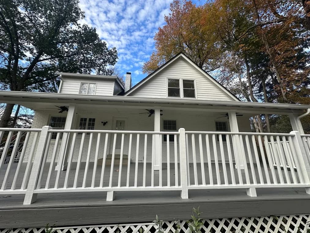 This is an image from Alarcon Pro Painting & Design, located in North Carolina. The exterior photo shows a freshly painted white two-story home with a covered front porch, white railing, and gray deck surrounded by autumn trees.