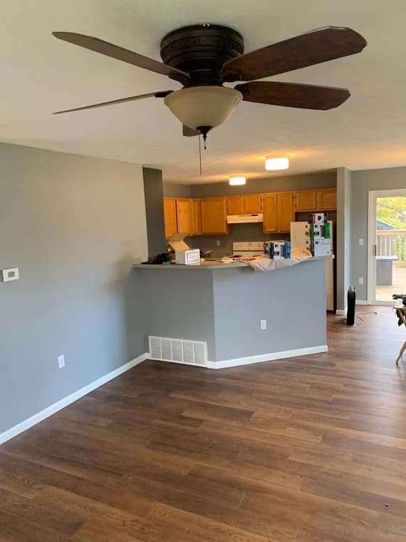 This is an image from Staib & Son Painting & Decorating LLC company, located in Jackson, MI The image is a freshly painted open-concept living room and kitchen with light gray walls, white trim, dark wood floors, ceiling fan, and natural wood kitchen cabinets.