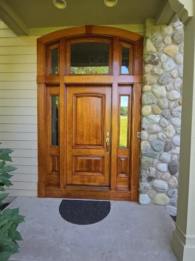 This is an image from Level Up Painting, located in New York. The photo shows a refinished wooden front door with a rich warm stain, framed by stonework and surrounded by narrow side windows.