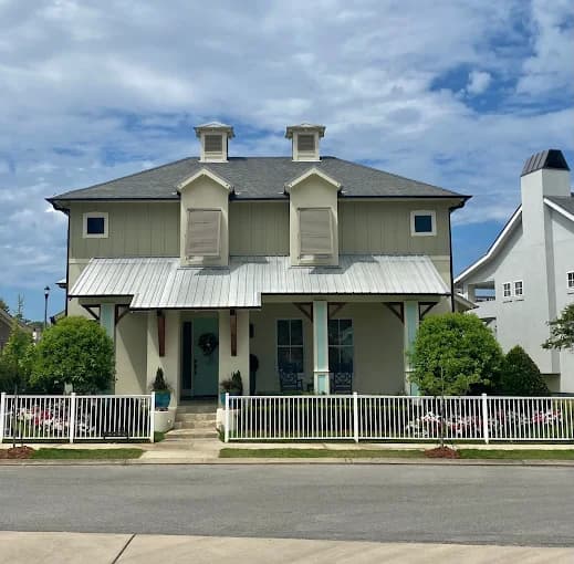 This is an image from Southern Dropcloth Painting LLC, located in Louisiana. The image shows a freshly painted two-story home with a light green exterior, white trim, and a metal roof, surrounded by a white picket fence and well-kept landscaping.