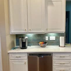 This is an image from Pro Edge Painting, showing a kitchen area with white cabinetry, a stainless steel dishwasher, and a light green tile backsplash. A Keurig coffee maker and a bowl of fruit are placed on the counter.