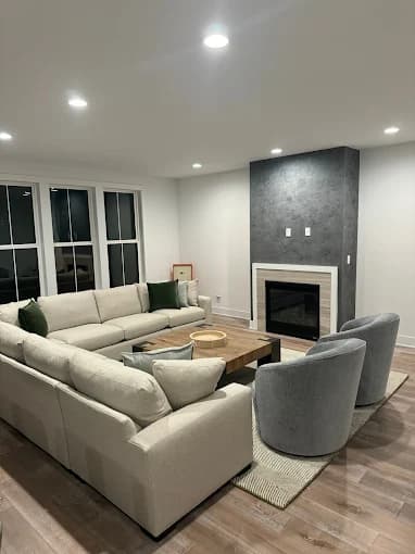 This is an image from Jay's Pro Painting, located in New York. The modern living room features white walls, recessed lighting, a sectional sofa, two gray chairs, and a fireplace with a textured gray accent wall.