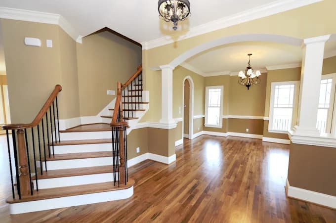 This is an image from EchoHousePainting, located in Massachusetts. The photo shows a freshly painted interior featuring warm beige and white walls, elegant crown molding, and polished hardwood floors surrounding a staircase with wrought-iron railings.