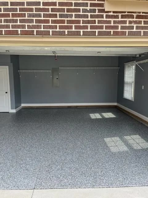 This is an image from Triple G Custom Painting Drywall & Roofing LLC, located in Indiana. The image shows a clean, newly coated garage with gray epoxy flooring and light gray walls, reflecting a polished and durable finish.