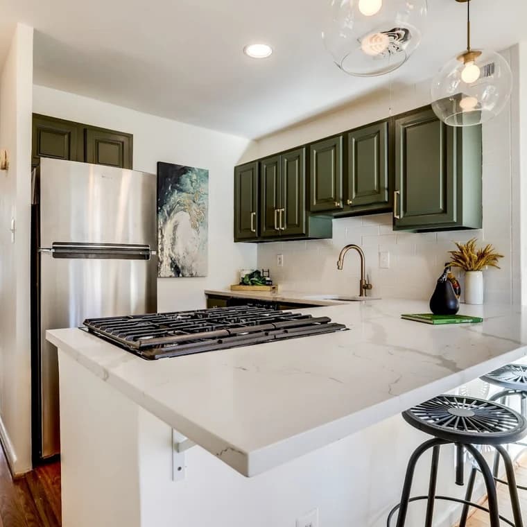 This is an image from Klappenberger & Son, located in Maryland. The photo shows a modern kitchen with dark green cabinets, marble countertops, stainless steel appliances, and industrial barstools.