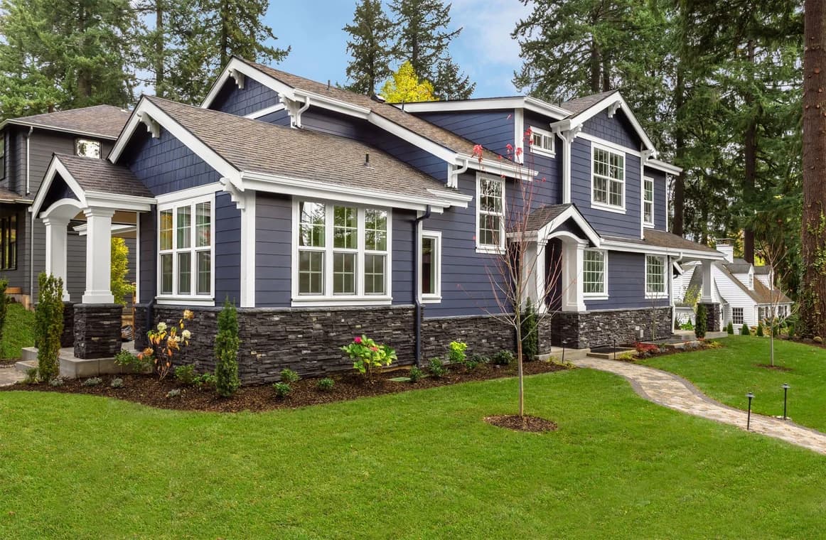 Exterior of a two-story home with navy blue siding, white trim, and stone detailing along the lower half, surrounded by a green lawn and landscaping.
