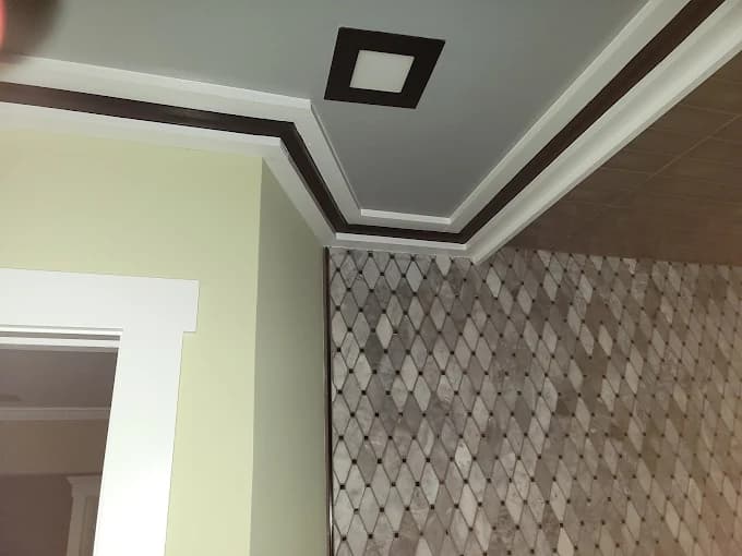 This is an image from Pro Edge Painting, showing a detailed ceiling corner with decorative trim painted in white, gray, and dark brown tones. The wall below features a patterned tile finish that adds texture to the space.