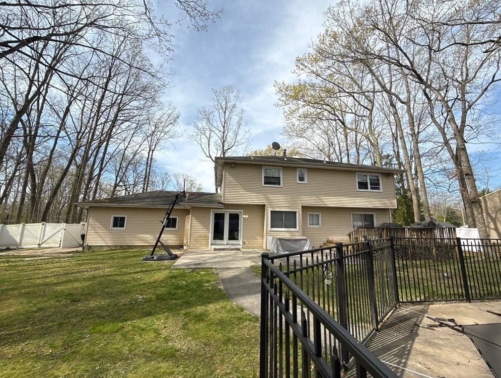 A beige two-story house with siding, located in a wooded backyard with a fenced-in area and basketball hoop.