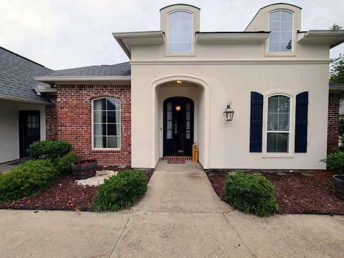 This is an image from Ozuna’s Painting Inc., located in Louisiana. The photo shows the freshly painted exterior of a modern home with cream-colored stucco walls, navy blue shutters, and a brick accent, highlighting the company’s professional exterior painting work.