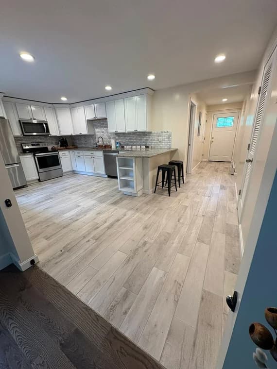 A renovated kitchen interior with new white cabinets, stainless steel appliances, and light gray tile flooring, updated by Titanium Painting & Renovations. The space has recessed lighting and a modern open layout.