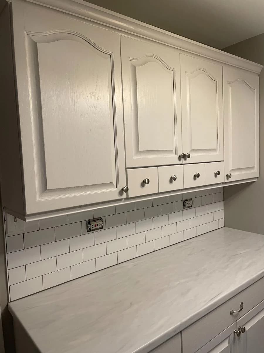 This is an image from A Plus Decorating Inc., located in Illinois. The image shows a kitchen cabinet painting project with white painted upper cabinets, subway tile backsplash, light countertop, and gray surrounding walls.