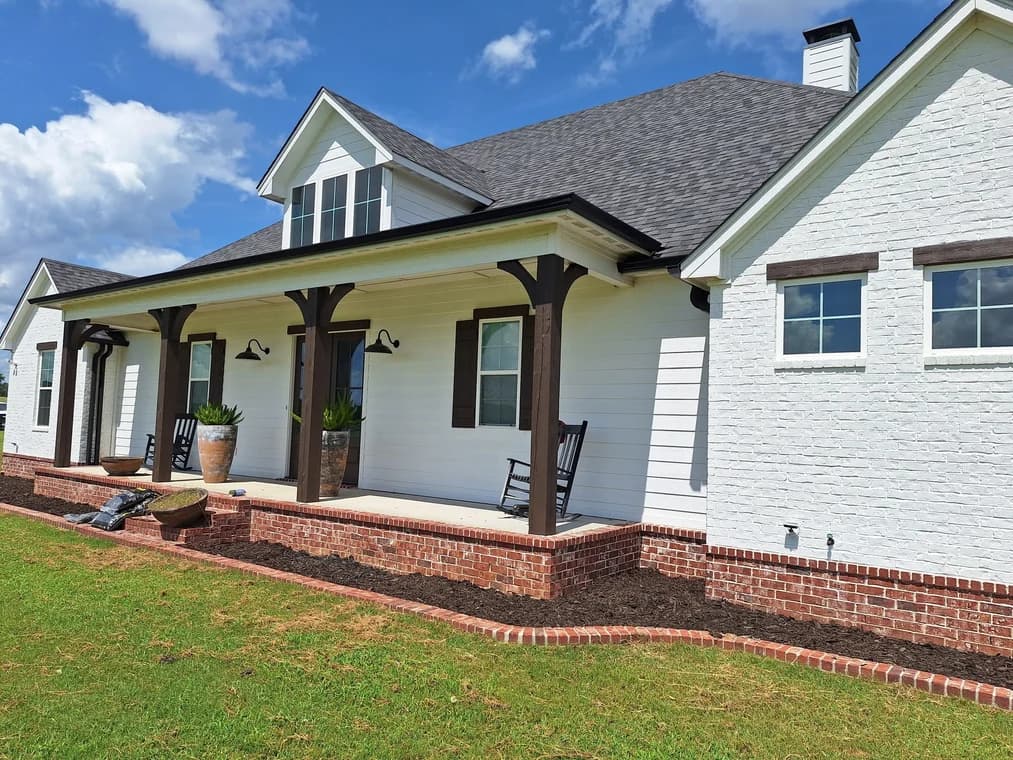 "This is an image from Reeves Painting, located in Texas. The exterior view shows a white brick home with dark wood porch posts, black shutters, and landscaped garden beds."