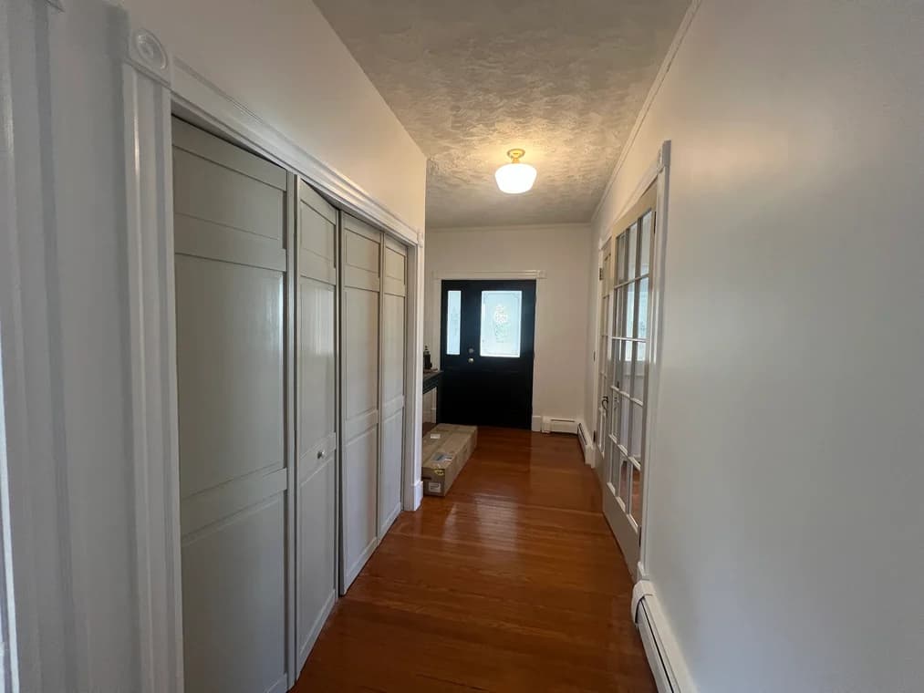 This is an interior hallway painted by Rodrigo's Painting. It shows white walls, freshly painted trim, wooden floors, and closet doors leading toward a black front door with a decorative glass insert.