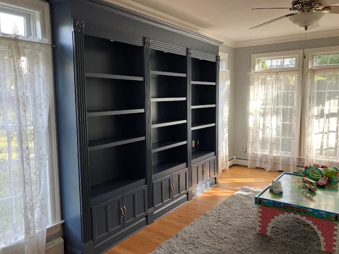 This is an image from Anthony's Painting & Decorating, Inc., located in New York. The photo shows a built-in wall unit painted in a dark charcoal finish with detailed trim, lower cabinet doors, and open shelving in a bright sunlit room.