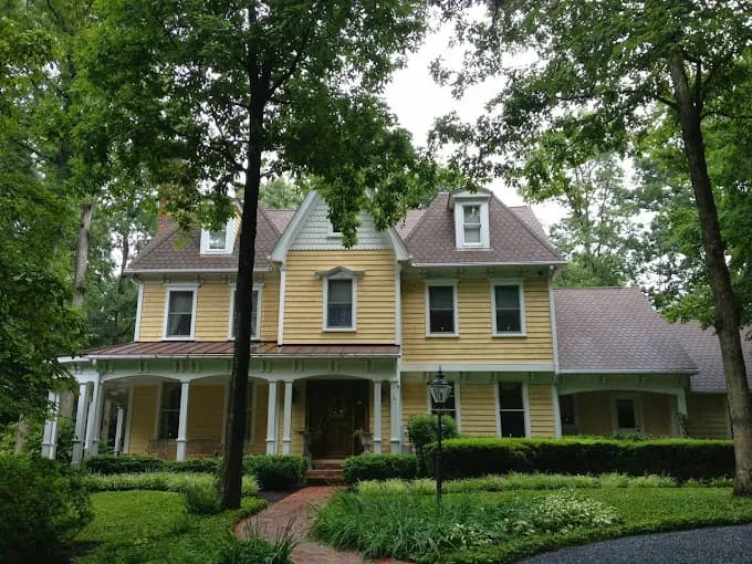 This is an image from CertaPro Painters of Columbia, located in Maryland. The image shows a large two-story yellow home with white trim, surrounded by tall trees and lush greenery.