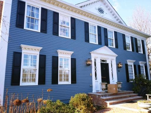 Exterior of a large colonial-style home with blue siding, white trim, and black shutters, freshly painted by Touch of the Brush Painting.