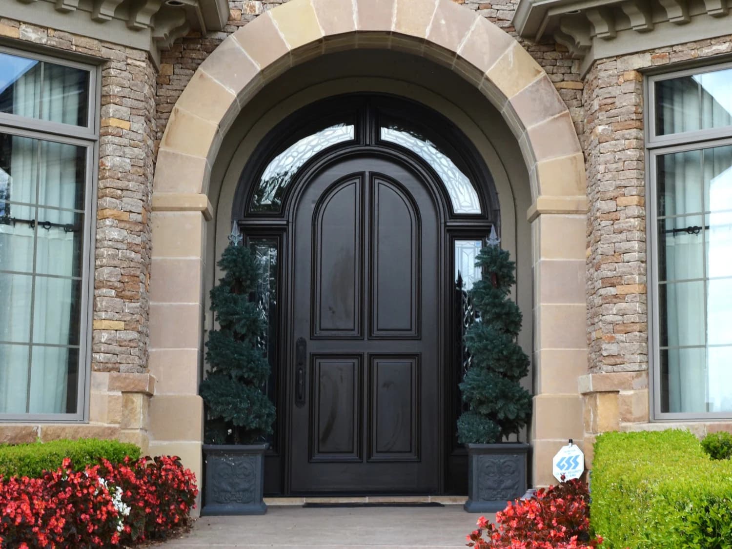 This is an image from JT Paint & Design, located in Oklahoma. The photo shows a beautifully painted dark wood front door framed by stonework, with decorative plants and flowers enhancing the elegant home entrance.