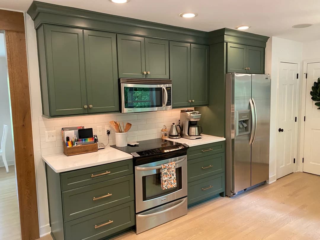 This is an image from Joyful Cabinet Painting, located in Ohio. The image shows a finished kitchen with dark green painted cabinets, modern hardware, white subway tile backsplash, stainless steel appliances, and hardwood flooring, highlighting high-end cabinet refinishing and interior painting services.