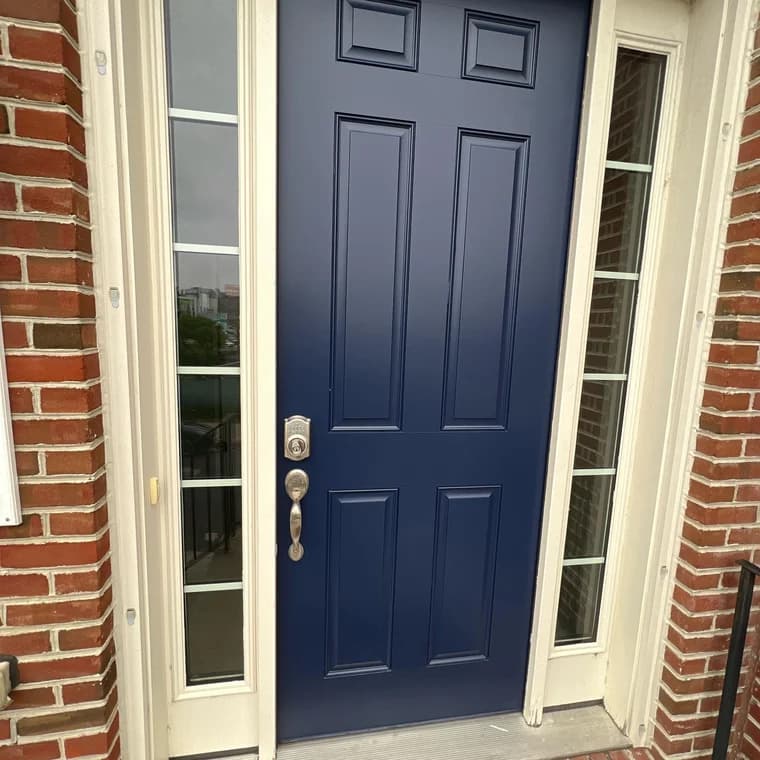 This is an image from Paint Perfection, located in Maryland. The close-up shot displays a newly painted navy blue front door framed by white trim and sidelights against a red brick exterior.