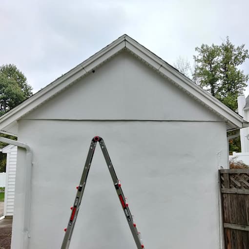 This is an image from Pro Painters Plus LLC company, located in Perkasie, PA. The image is a freshly painted white exterior of a detached garage with a clean gable roof, a wooden fence to the side, and an aluminum ladder positioned in front.