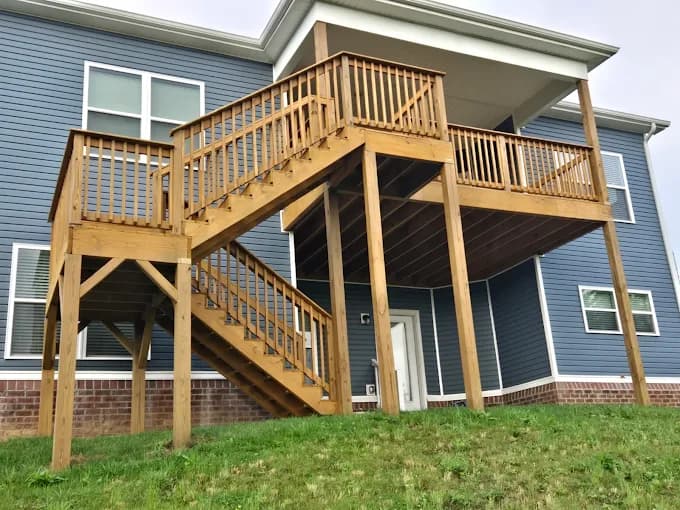 This is an image from Quick Painting LLC, located in Kentucky. The image shows a newly built natural wood deck with stairs and railings attached to a blue-sided home, showcasing clean construction and exterior finishing.