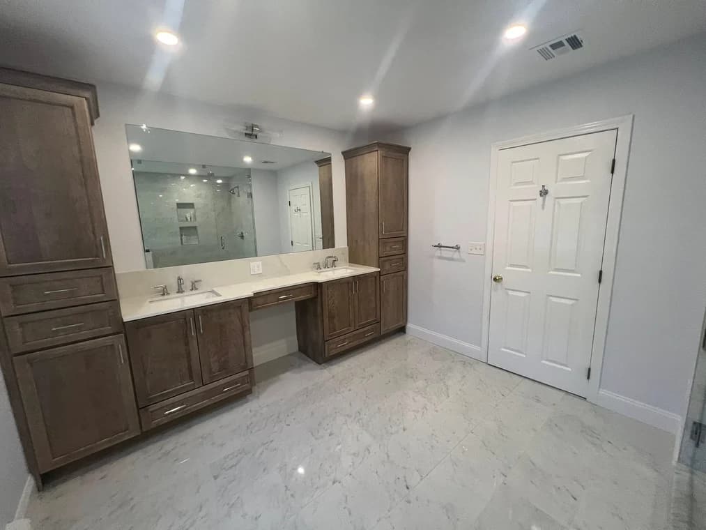 This is an image from Powell Painting & Home Services, located in Massachusetts. The photo shows a modern bathroom with freshly painted light gray walls, dual sinks with wood cabinetry, marble tile flooring, and a large mirror above the vanity