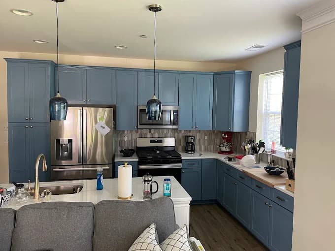 This is an image from Firefly Painting Inc, located in Colorado. The kitchen features newly painted blue cabinets, stainless steel appliances, pendant lighting, and a white countertop.