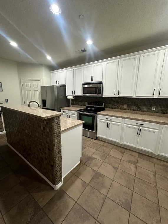 This is an image from CertaPro Painters of Albuquerque, located in New Mexico. The photo shows a modern kitchen with freshly painted white cabinets, stainless steel appliances, and a brown mosaic tile backsplash.