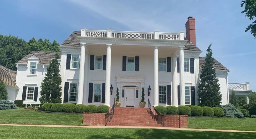 This is an image from Greg Baker Painting. The image shows an exterior colonial-style home in Kansas City KS with white painted siding, black shutters, and a freshly painted brick entrance.