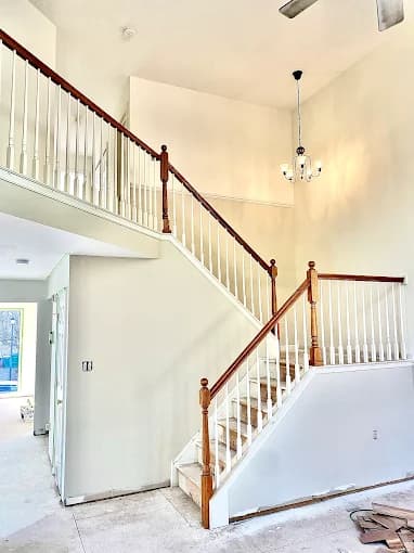This is an image from Pro Painters Plus LLC company, located in Perkasie, PA. The image is a freshly painted interior stairway with white walls, wooden handrails, white spindles, and a modern chandelier in a two-story foyer with natural light.