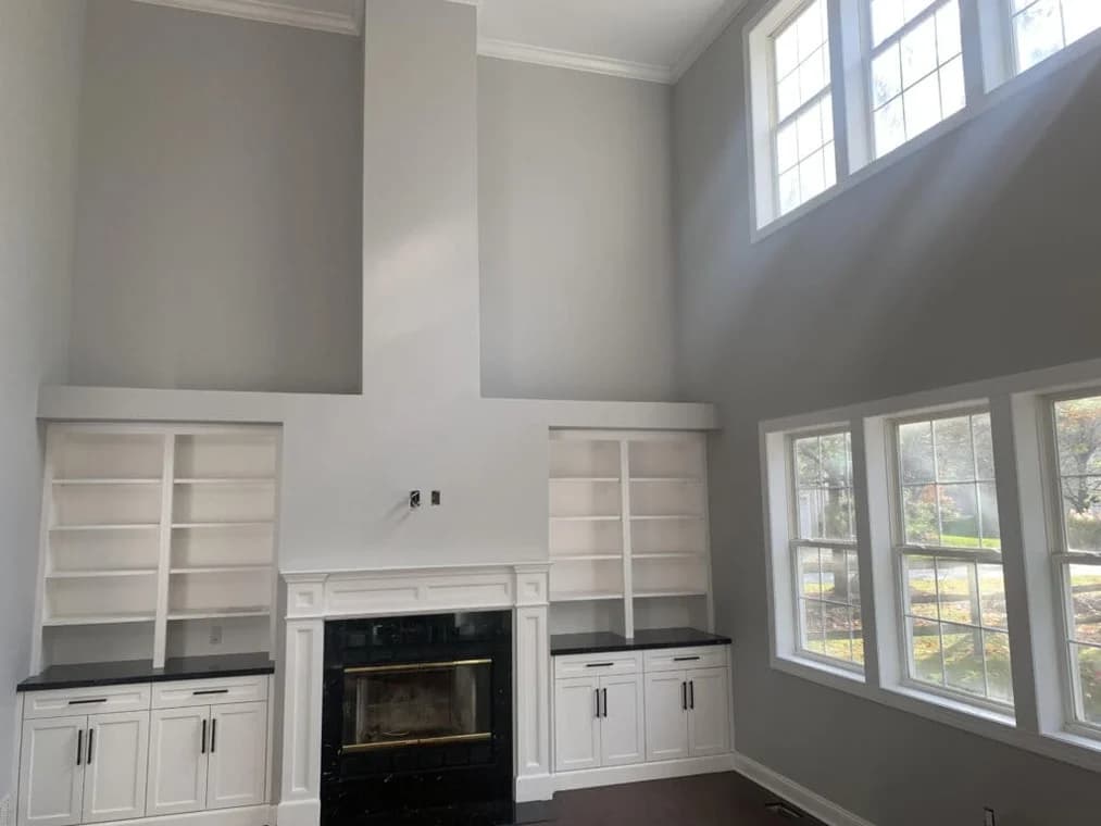 This is an image from Hoffman Interior Painting, located in Michigan. The image shows a freshly painted living room with tall vaulted ceilings, built-in white shelving, large windows, and soft gray walls surrounding a fireplace demonstrating professional interior wall and trim painting services.
