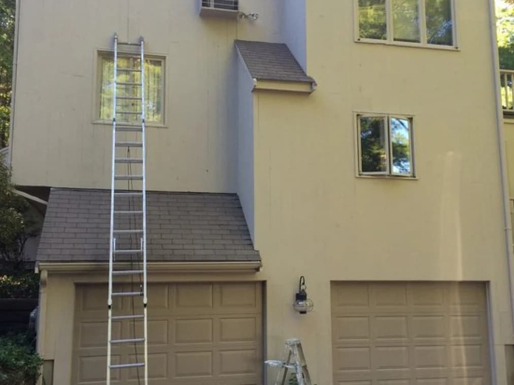 This is an image from Nelson Painting, Inc., located in Massachusetts. It shows a tall beige house with garage doors, ladders set up, and preparation underway for exterior painting.