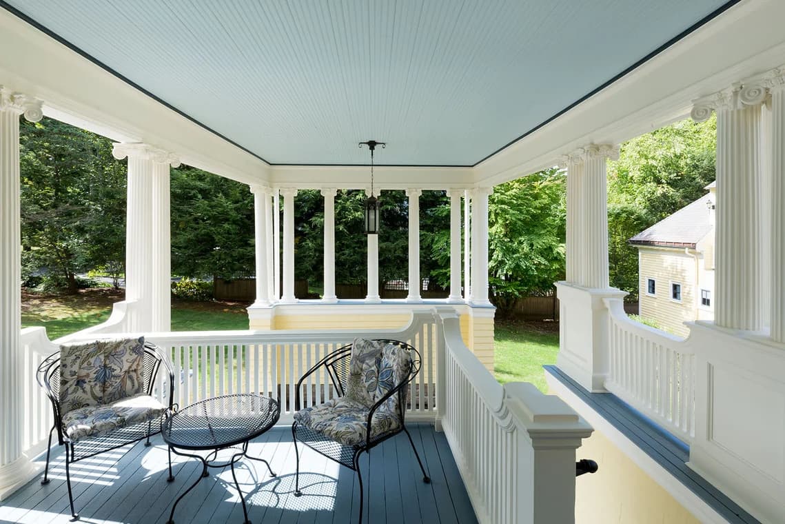 This is an image from Catchlight Painting, located in Massachusetts. It shows a detailed porch with white columns and railings, a blue ceiling, and outdoor seating.