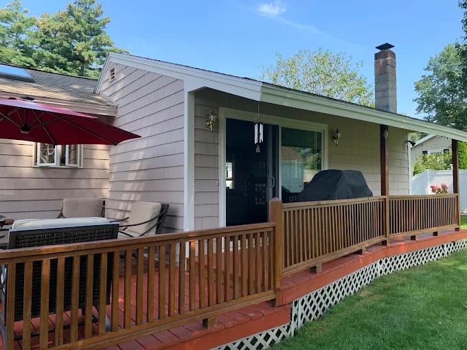 This is an image from Castle Complements Painting Co., located in Massachusetts. It features the back patio of a home with beige siding, a wooden railing, and a red-stained deck floor.