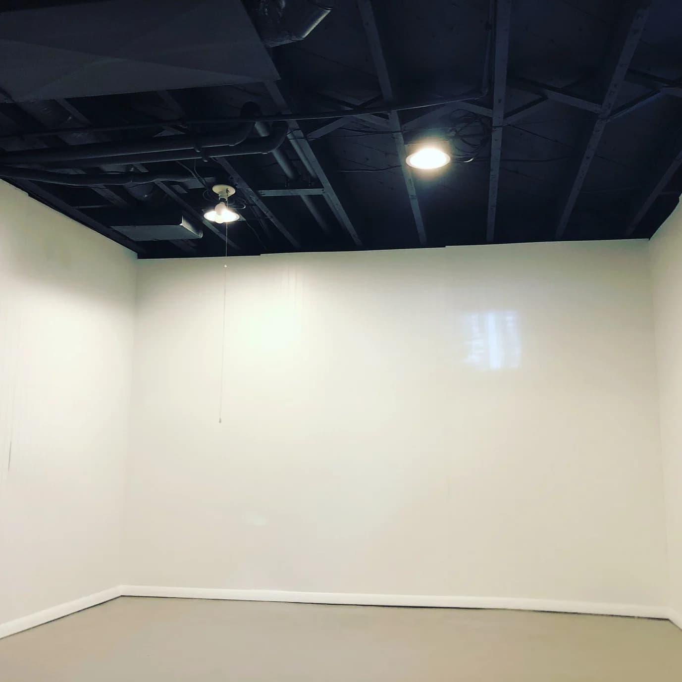 This is an image from Graystone Painting and Refinishing. The image is an interior basement room in Kansas City KS with white painted walls, exposed black ceiling joists, and overhead lighting.