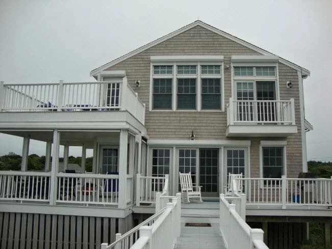 This is an image from CertaPro Painters of Cape Cod, located in Massachusetts. The photo shows a two-story coastal home with fresh gray shingle siding and crisp white trim, railings, and decks, highlighting a clean and professional exterior paint job.