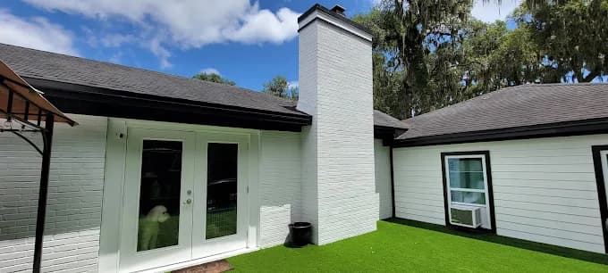This is an image from Rockstar Painting, Inc. company, located inJacksonville, FL. The image is a modern white-painted brick home exterior with black trim and artificial turf, featuring French doors and central chimney, painted by Rockstar Painting Inc.