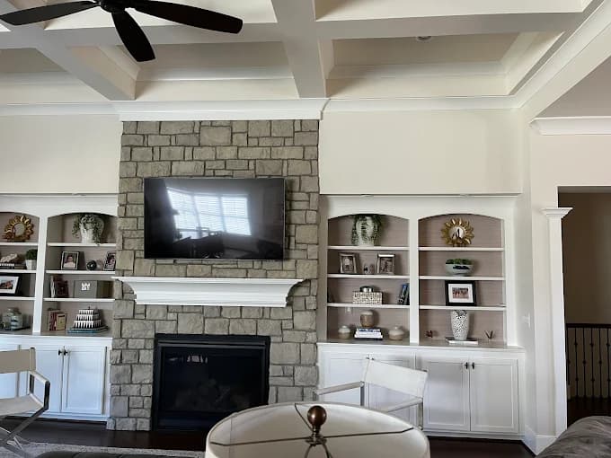 This is an image from Serious Business Painting LLC, located in Kentucky. The image shows a professionally painted living room with a stone fireplace, white built-in shelving, coffered ceiling, and smooth neutral wall colors.