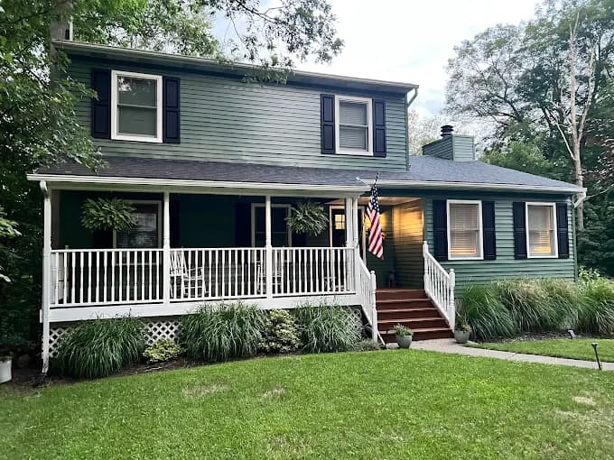 This is an image from Willow Tree Painting & Home Improvement LLC, located in New York. The photo shows a two-story green home with black shutters, a white wraparound porch, and warm entry lighting during the early evening.